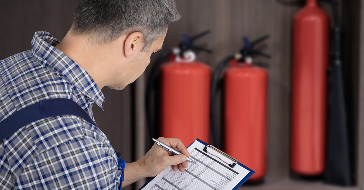 A worker inspects an emergency light fixture with a tablet, performing a fire safety check.