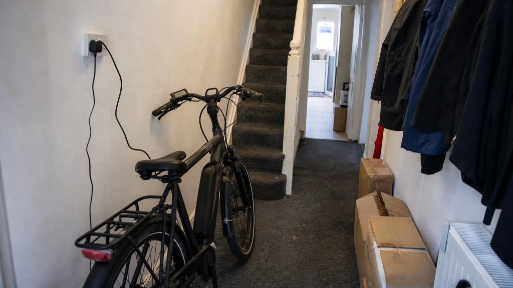 An e-bike charging in a narrow HMO hallway beside the stairs with coats and boxes nearby
