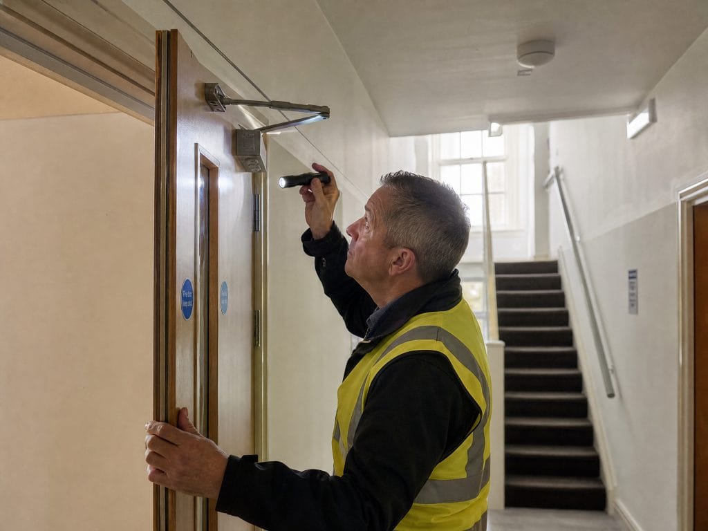 A fire risk assessor checks a fire door self-closer and seal in the shared hallway of an HMO, with communal stairs behind